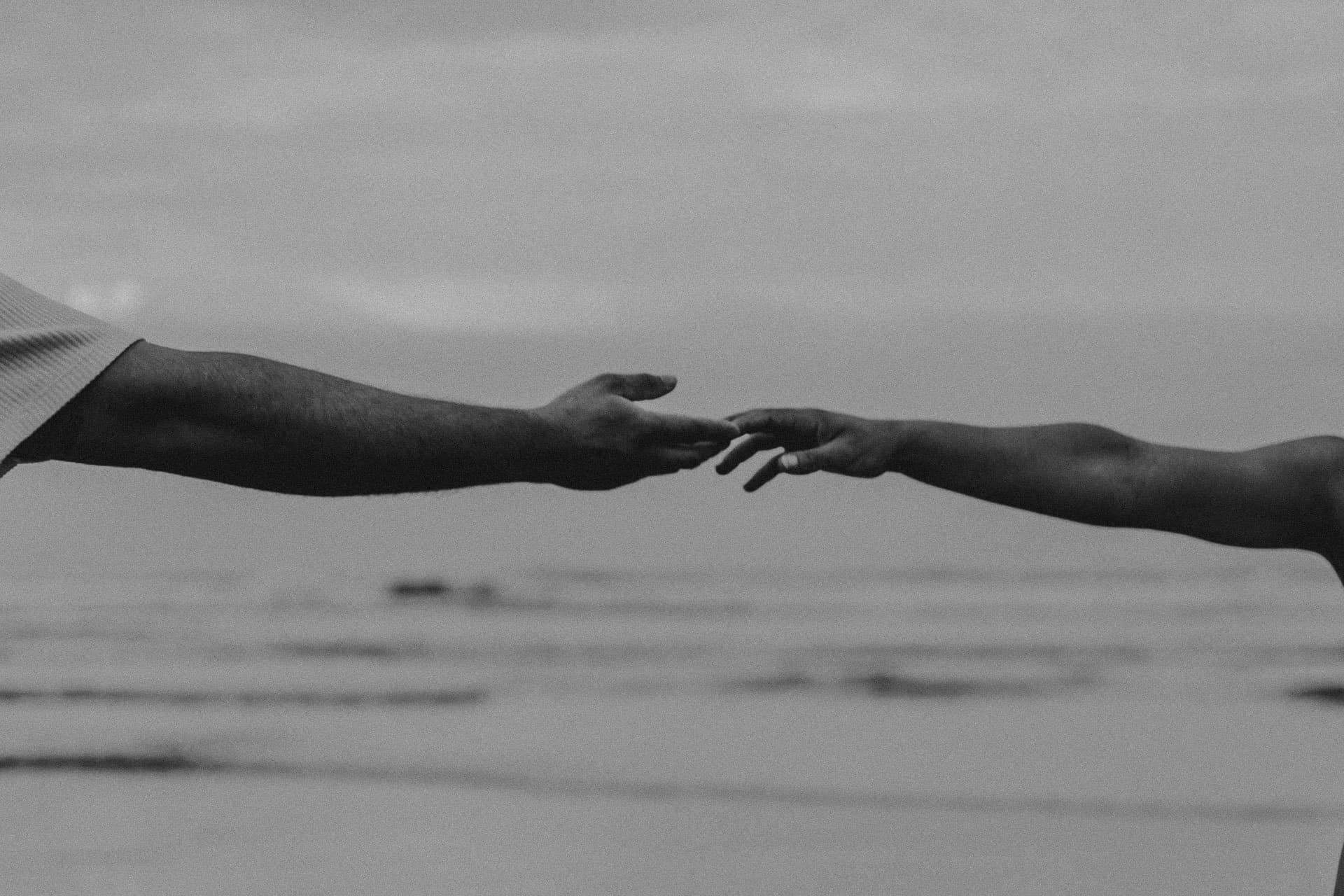 Dois braços se aproximando sobre a praia, pontas dos dedos quase se tocando